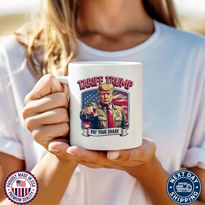 May include: White ceramic coffee mug with a graphic of Donald Trump in front of an American flag. The text "Tarife Trump" and "Pay Your Share" are printed on the mug.