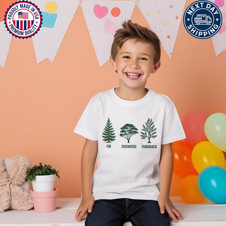 Pu&ograve; includere: Una maglietta bianca con illustrazioni di alberi verdi e le parole "FIR", "DOGWOOD" e "TAMARACK". Un bambino indossa la maglietta e sorride. L'immagine include anche loghi patriottici e relativi alla spedizione.