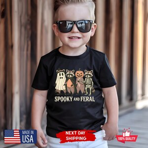 Puede incluir: Camiseta negra con un gr&aacute;fico de Halloween que presenta mapaches disfrazados de fantasma, vampiro, calabaza y esqueleto. El texto "SPOOKY AND FERAL" est&aacute; debajo. La camiseta la lleva un ni&ntilde;o con gafas de sol.