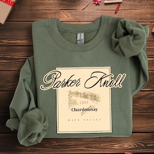 Può includere: Felpa verde salvia con un design di etichetta di vino. L'etichetta recita "Parker Knoll" in elegante corsivo, con "1993 Chardonnay Napa Valley" sotto un'illustrazione di vigneto. La felpa è realizzata in materiale morbido.