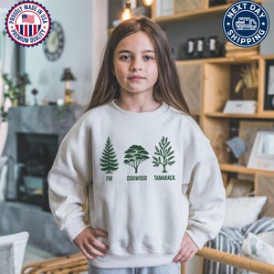 Pu&ograve; includere: Una felpa color crema con tre illustrazioni di alberi verdi e le parole "FIR", "DOGWOOD" e "TAMARACK" stampate sotto. La felpa ha un girocollo e maniche lunghe. L'immagine include anche un sigillo "Proudly Made in USA" e un logo "Next Day Shipping".
