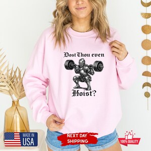 May include: Light pink sweatshirt with a graphic of a knight lifting weights, and the text "Dost Thou even Hoist?" in black. The design is centered on the front of the long-sleeved crew neck top.