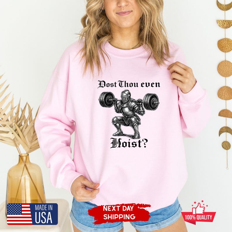 May include: Light pink sweatshirt with a graphic of a knight lifting weights, and the text "Dost Thou even Hoist?" in black. The design is centered on the front of the long-sleeved crew neck top.