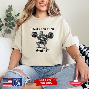 May include: Beige t-shirt with a black and white graphic of a knight lifting weights, with the text "Dost Thou even Hoist?" in a gothic font. The shirt is made of a soft material.