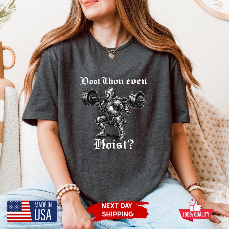 May include: Dark gray t-shirt featuring a graphic of a knight lifting weights with the text "Dost Thou even Hoist?" in a medieval font. The shirt has short sleeves and a crew neck. The image also includes "Made in USA" and "Next Day Shipping" text.
