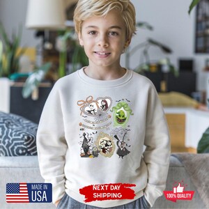 Może przedstawiać: Kremowa bluza z postaciami z kresk&oacute;wek w ramkach z filmu o tematyce Halloween. Projekt zawiera Jacka Skellingtona, Sally i inne postacie. Bluza ma napis "MADE IN USA" i "NEXT DAY SHIPPING".