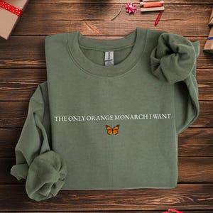 May include: Olive green sweatshirt with the text "THE ONLY ORANGE MONARCH I WANT" and a small orange butterfly graphic. The sweatshirt has a crew neck and long sleeves.