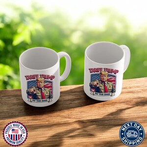 May include: Two white ceramic mugs with a graphic of Donald Trump in front of an American flag. The text on the mug reads "Tariff Trump Pay Your Share".