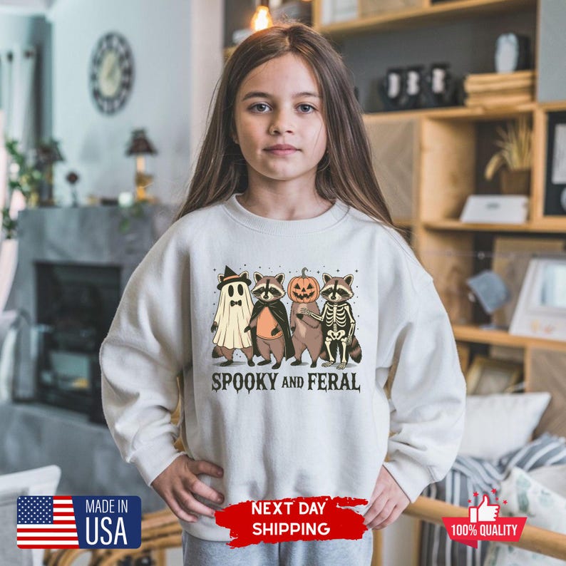 Puede incluir: Sudadera crema con un dise&ntilde;o de Halloween que presenta un fantasma, mapaches y una calabaza. El texto "SPOOKY AND FERAL" est&aacute; impreso debajo. Hecho en los EE. UU.