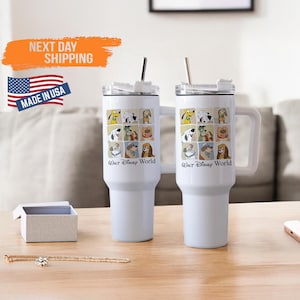 Puede incluir: Dos vasos aislados blancos con asas y pajitas de acero inoxidable. Cada vaso presenta un collage de personajes de perros de dibujos animados y el texto "Walt Disney World". Los vasos están sobre una superficie de madera.