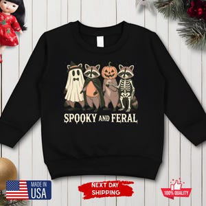 Puede incluir: Sudadera negra de manga larga con un dise&ntilde;o de Halloween. El dise&ntilde;o muestra cuatro mapaches disfrazados de fantasma, vampiro, calabaza y esqueleto. El texto "SPOOKY AND FERAL" est&aacute; debajo.