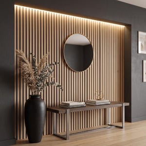 Op de afbeelding: Een wand met verticale lichte houten latten en geïntegreerde verlichting, een ronde spiegel, een donkerbruine houten bank met metalen poten en een hoge zwarte vaas met gedroogd pampasgras en eucalyptusstengels.