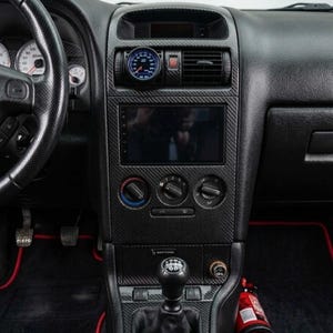 Könnte beinhalten: Eine Auto-Mittelkonsole mit Carbonfaser-Muster, einem zentralen Touchscreen-Display, drei runden Klimaregelungsknöpfen, einem manuellen Schalthebel und einer runden Anzeige über den Lüftungsschlitzen mit dem Schriftzug Bertone.
