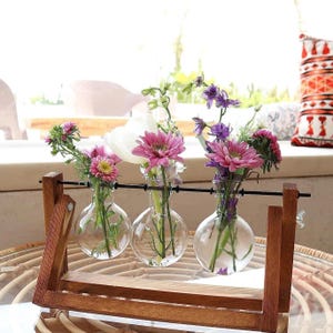 May include: A wooden flower vase display with three clear glass globe vases, each holding fresh flowers in shades of pink, purple, and white. The wooden frame has a dark finish and a black metal rod.