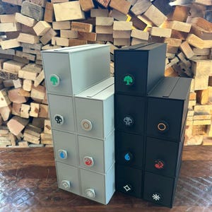 May include: Two vertical storage organizers with multiple drawers, one in light gray and one in black. Each drawer features a circular knob with a distinct colored icon representing different elements, including a tree, skull, sun, flame, and water drop.