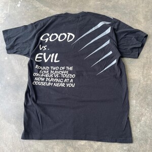 May include: A black short-sleeve t-shirt featuring white text that reads GOOD VS. EVIL, ROUND TWO OF THE ECHL PLAYOFFS COLUMBUS VS. TOLEDO NOW PLAYING AT A COLISEUM NEAR YOU, accompanied by a series of five diagonal silver claw mark graphics.