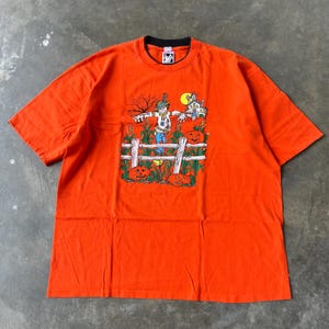 May include: Orange t-shirt with a Halloween-themed graphic. The graphic features a scarecrow, pumpkins, a fence, and a haunted house. The shirt has a black collar and short sleeves. The shirt is made of a soft material.