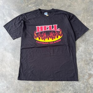 May include: Black t-shirt featuring the word "HELL" in red block letters above a yellow and red flame design. Below the flames, the text "CAYMAN ISLANDS" is printed in red. The shirt is made of a soft material.