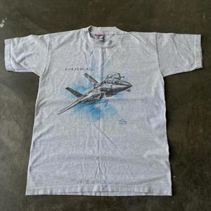 May include: A light gray t-shirt featuring a detailed illustration of an F-14 Tomcat fighter jet. The graphic includes the text "F-14 TOMCAT" above the jet and a blue cloud-like background. The shirt has a crew neck and short sleeves.