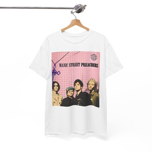 May include: White t-shirt featuring a pink background with the text "MANIC STREET PREACHERS" and a photo of the band. The shirt is hanging on a wooden hanger.
