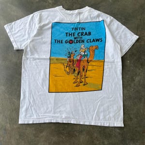 May include: A white short-sleeve t-shirt featuring a rectangular graphic on the back. The print depicts a desert scene with two camels and the text TINTIN THE CRAB WITH THE GOLDEN CLAWS, showing characters riding through a yellow sand landscape.