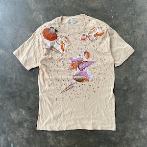 May include: Tan short-sleeved t-shirt featuring a colorful seashell design. The design includes various seashells in shades of orange, purple, and white, with small confetti-like details scattered across the shirt. The shirt is made of a soft, comfortable material.