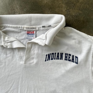 May include: White polo shirt with a collar and buttoned placket. The shirt features the text "INDIAN HEAD" in navy blue block letters. The tag reads "RACHEL HOCHMAN" and "ANVIL".