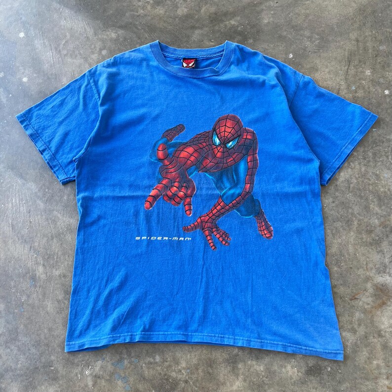 Pode incluir: Uma t-shirt azul brilhante de manga curta com um gr&aacute;fico da personagem Spider-Man numa posi&ccedil;&atilde;o agachada. A personagem &eacute; vermelha e azul com um padr&atilde;o de teia. O texto Spider-Man est&aacute; impresso a branco por baixo do gr&aacute;fico na frente.