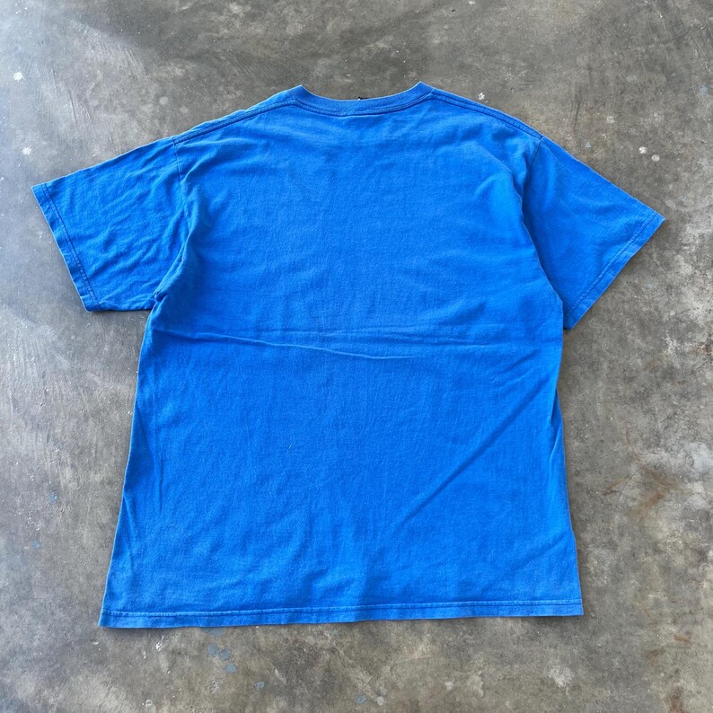 Pode incluir: Uma t-shirt de manga curta de cor azul brilhante s&oacute;lida com gola redonda. A pe&ccedil;a &eacute; exibida estendida, revelando a parte de tr&aacute;s da t-shirt, que apresenta uma superf&iacute;cie de tecido lisa sem padr&otilde;es com costuras padr&atilde;o nas mangas e na bainha.