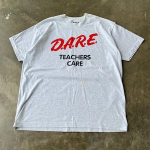 May include: A light gray t-shirt with the text "D.A.R.E. TEACHERS CARE" in red and black. The shirt is made of a soft material and has short sleeves. The t-shirt is a vintage item.