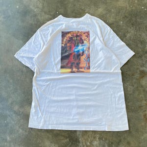 May include: White t-shirt with a graphic on the back featuring a figure with blue energy and the text "Apocalypse Cheat". The graphic is in shades of red, blue, and yellow. The shirt is made of a soft, white fabric.