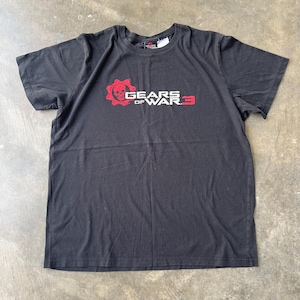 May include: Black t-shirt featuring the Gears of War 3 logo in red and white. The logo includes a skull within a cog design. The shirt is short-sleeved and appears to be made of cotton.