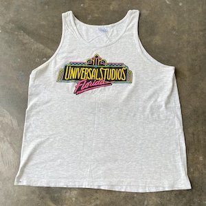 May include: A light gray tank top featuring the Universal Studios Florida logo in a neon design. The logo includes the words "Universal Studios" in yellow and "Florida" in pink, with a black outline. The tank top is made of a soft, lightweight material.