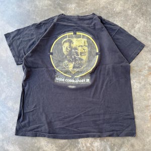 May include: Black t-shirt featuring a circular graphic from Wing Commander III. The graphic includes images of characters and the text "Wing Commander III: Heart of the Tiger". The shirt is made of a soft material.