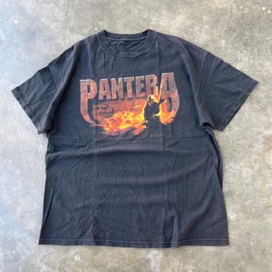 May include: Black t-shirt featuring the band name "PANTERA" in a distressed orange and red font. The graphic includes a silhouette of a person and flames. The text "Brother Dime" is below the graphic.