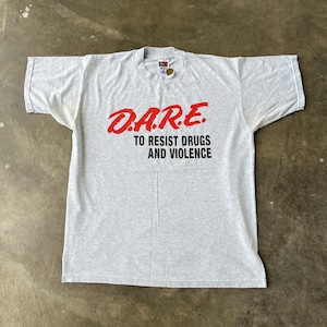 May include: A light gray t-shirt with the text "D.A.R.E. TO RESIST DRUGS AND VIOLENCE" in red and black. The shirt is laid flat on a concrete surface.
