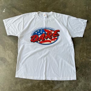 May include: White t-shirt featuring the San Ramon Police Department D.A.R.E. logo. The logo is an oval design with a red and white checkered pattern, blue stars, and the text "DARE" in red and white.