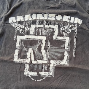 Pu&ograve; includere: T-shirt grigio scuro con il nome della band "RAMMSTEIN" sopra un design a croce stilizzato bianco con catene. La grafica &egrave; bianca e ha un aspetto vintage usurato. La maglietta &egrave; realizzata in tessuto morbido e usurato.