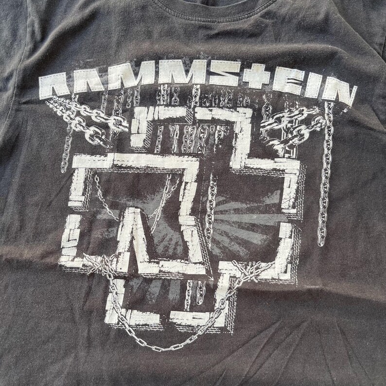 Pu&ograve; includere: T-shirt grigio scuro con il nome della band "RAMMSTEIN" sopra un design a croce stilizzato bianco con catene. La grafica &egrave; bianca e ha un aspetto vintage usurato. La maglietta &egrave; realizzata in tessuto morbido e usurato.