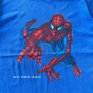 Pode incluir: Uma t-shirt azul brilhante com um gr&aacute;fico grande da personagem Spider-Man numa pose agachada com padr&otilde;es de teia vermelhos e pretos. A personagem tem olhos brancos grandes com reflexos azuis. O texto SPIDER-MAN est&aacute; impresso em branco abaixo.
