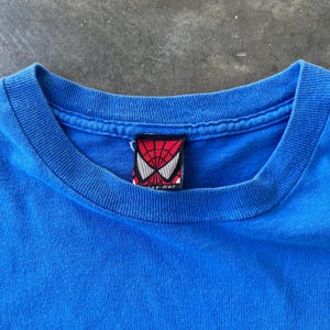 Pode incluir: Uma t-shirt azul brilhante com gola redonda canelada. No interior da gola, encontra-se uma etiqueta de tecido retangular com um padr&atilde;o de teia de aranha vermelho e preto, formas de olhos brancas e o texto Spider-Man impresso a preto.