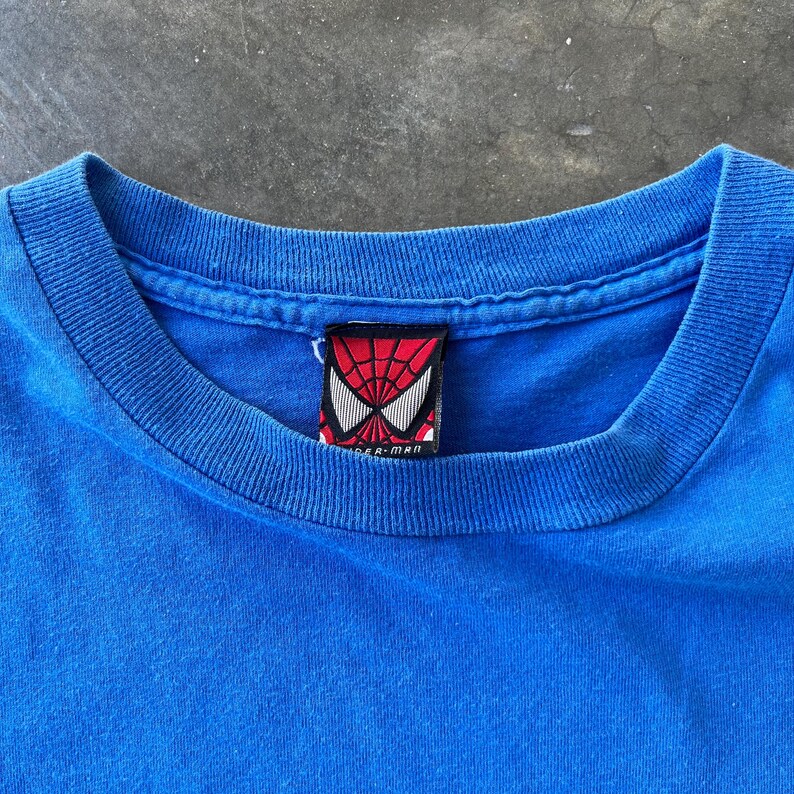 Pode incluir: Uma t-shirt azul brilhante com gola redonda canelada. No interior da gola, encontra-se uma etiqueta de tecido retangular com um padr&atilde;o de teia de aranha vermelho e preto, formas de olhos brancas e o texto Spider-Man impresso a preto.