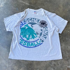 May include: A heather gray t-shirt featuring a large graphic of a German soccer emblem. The emblem includes a stylized eagle in teal and white, surrounded by text in blue and black. The shirt is laid flat.
