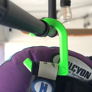 Puede incluir: Un colgador verde brillante para equipo de buceo está sujeto a una barra de metal negra. El colgador sostiene un chaleco de buceo morado y negro. El chaleco tiene una correa blanca y gris con un parche azul y blanco con la letra "H".