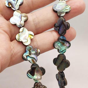 May include: A strand of clover-shaped beads crafted from iridescent abalone shell. Each bead displays a unique natural pattern with shifting hues of green, blue, purple, and silver, strung together to form a continuous decorative jewelry component.