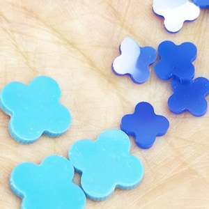 May include: A collection of small, four-leaf clover-shaped embellishments in varying shades of blue. The pieces include matte turquoise and glossy royal blue finishes, with some featuring reflective surfaces that catch the light.