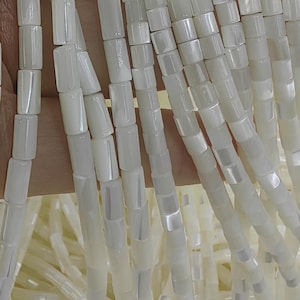 Peut inclure: Plusieurs brins de perles en nacre blanche disposés verticalement. Chaque perle présente une forme cylindrique lisse avec une finition polie et irisée qui reflète la lumière, créant un aspect cohérent et lumineux sur l'ensemble.