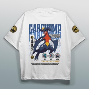 Op de afbeelding: Wit t-shirt met een Garchomp-afbeelding, een Dragon/Ground-type Pokémon, en Japanse tekst. Het ontwerp bevat blauwe, rode en gele accenten, met het nummer 445 en extra Pokémon-illustraties.