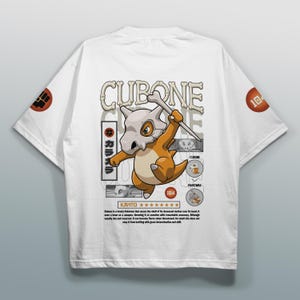 Puede incluir: Una camiseta blanca de manga corta con un gráfico del personaje Cubone sosteniendo un hueso. El diseño incluye el texto CUBONE, KANTO y párrafos descriptivos, junto con pequeños iconos circulares en las mangas y el área del pecho.