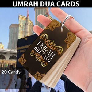 Puede incluir: Un juego de tarjetas Umrah Dua sostenido por una persona, con la Kaaba y un paisaje urbano de fondo. Las tarjetas son oscuras con detalles dorados y las palabras "Umrah Dua Card". El texto "20 Cards" es visible.
